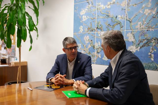 Dos hombres conversan en una oficina con arte en la pared y una planta al fondo.