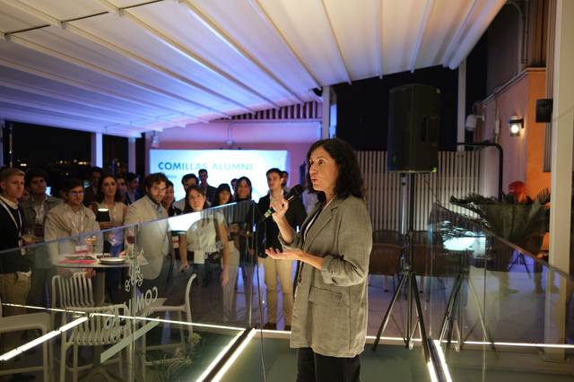 Una mujer habla frente a un grupo de personas en un evento al aire libre durante la noche.
