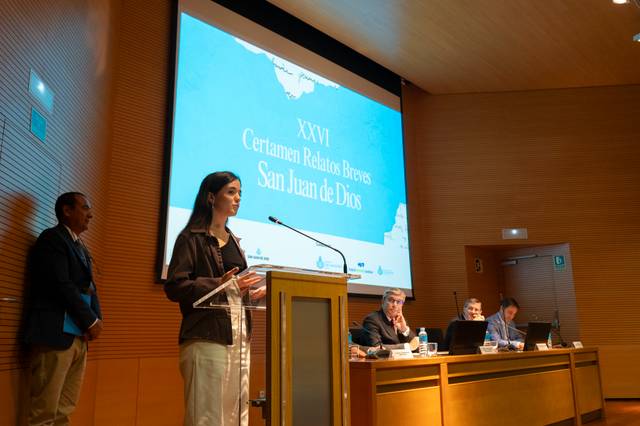Una mujer presenta un evento en un auditorio, mientras un grupo de personas la escucha desde una mesa.
