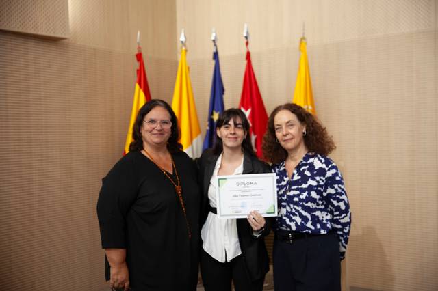 Tres mujeres posan junto a un diploma y banderas de fondo.