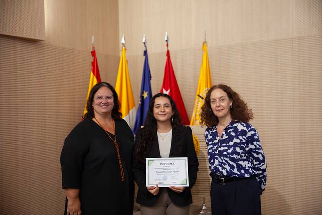 Tres mujeres posan en un evento de entrega de diplomas con banderas de fondo.