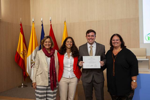 Cuatro personas están de pie en un evento, sosteniendo un certificado y rodeadas de banderas.