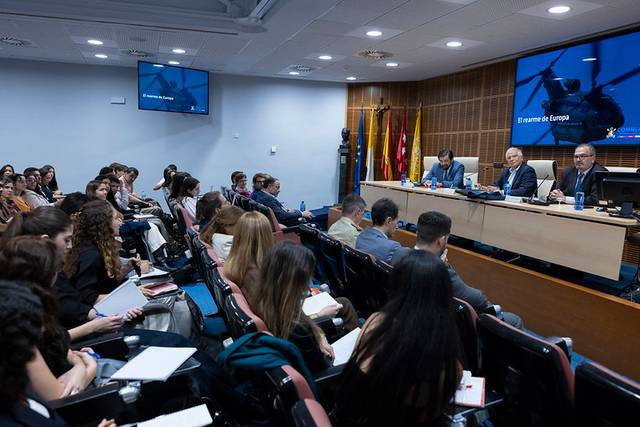 Un grupo de personas asiste a una conferencia en un salón con pantallas y banderas.