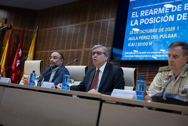 Un grupo de tres hombres discute en un evento sobre el rearme en una sala con carteles y banderas.