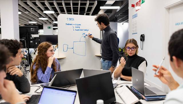 Un grupo de personas discute y trabaja en equipo en un ambiente de oficina mientras un integrante escribe en una pizarra.