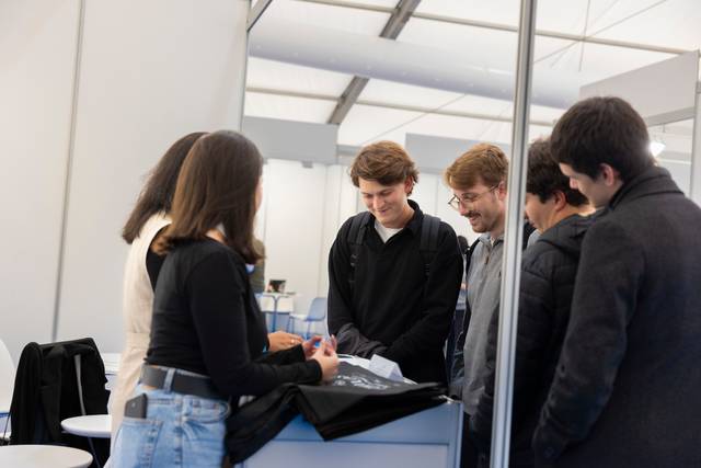 Un grupo de jóvenes interactuando en un evento al aire libre, observando un objeto sobre una mesa.