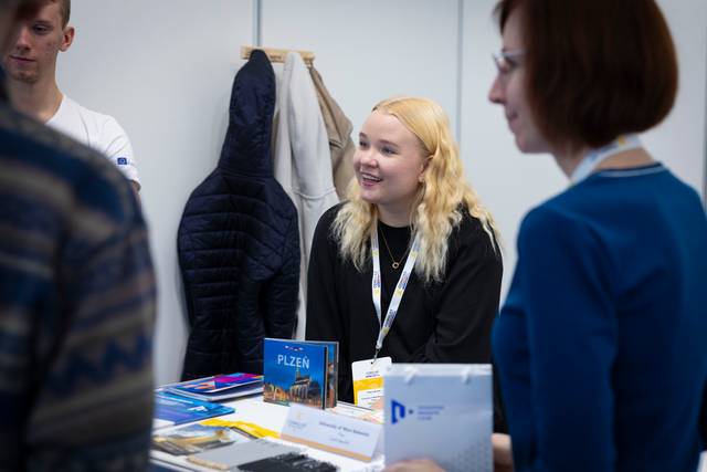 Una joven sonriente está atendiendo en un stand, rodeada de personas y materiales promocionales.