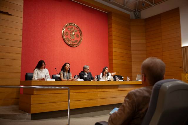 Una mesa de debate en una sala con personas sentadas y un público asistiendo.