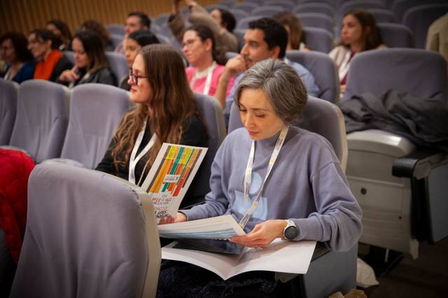 Una mujer revisa documentos mientras asiste a una conferencia con un público diverso detrás de ella.