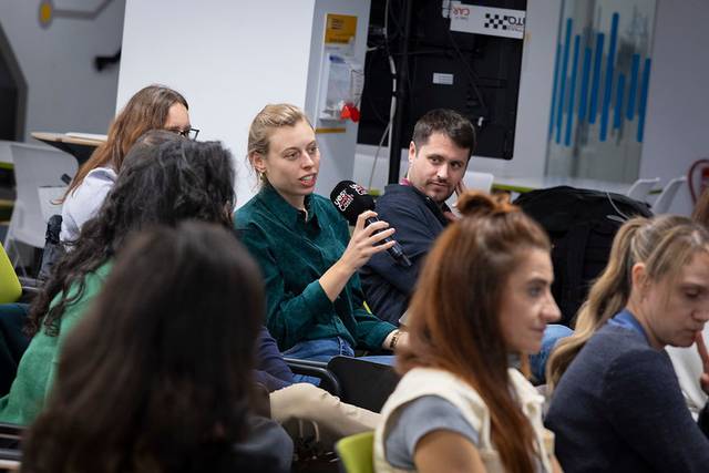 Un grupo de personas sentadas en un evento, donde una mujer está hablando con un micrófono.