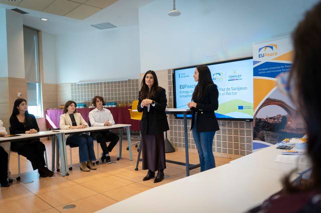 Una reunión con varias personas sentadas alrededor de una mesa donde dos mujeres están hablando frente a una pantalla de presentación.