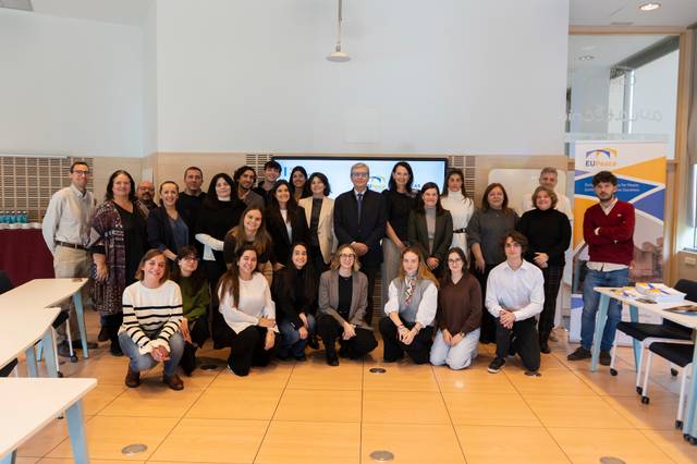 Un grupo de personas posando juntas en un salón con mesas y una presentación al fondo.