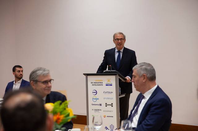 Un hombre está dando un discurso en un evento empresarial con varios asistentes escuchando atentamente.