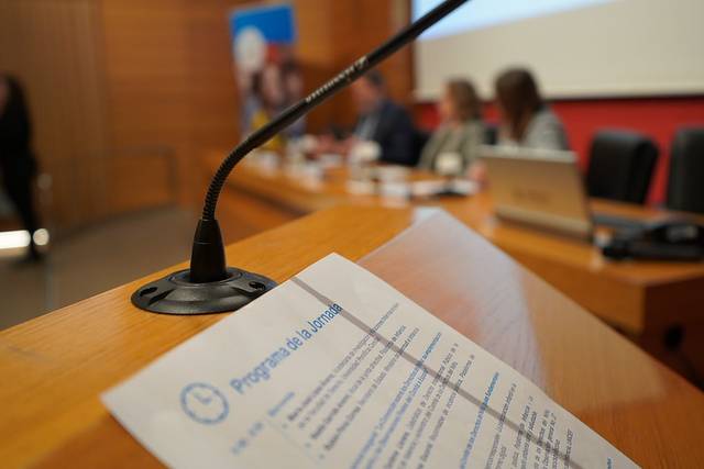 Una vista de un podio con un micrófono y un programa de la jornada en primer plano, con un panel de ponentes al fondo.