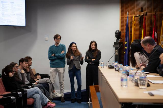 Un grupo de jóvenes presentan un proyecto frente a un auditorio, mientras un panel de expertos escucha atentamente.