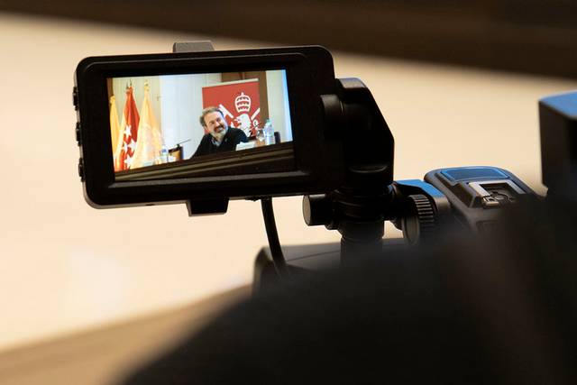 Una cámara graba a una persona hablando en una conferencia mientras se ven banderas en el fondo.
