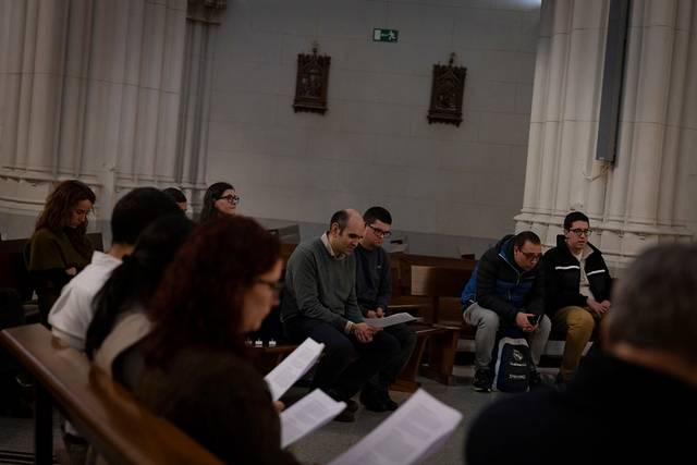 Un grupo de personas sentadas en un banco de una iglesia, leyendo documentos en un ambiente tranquilo.