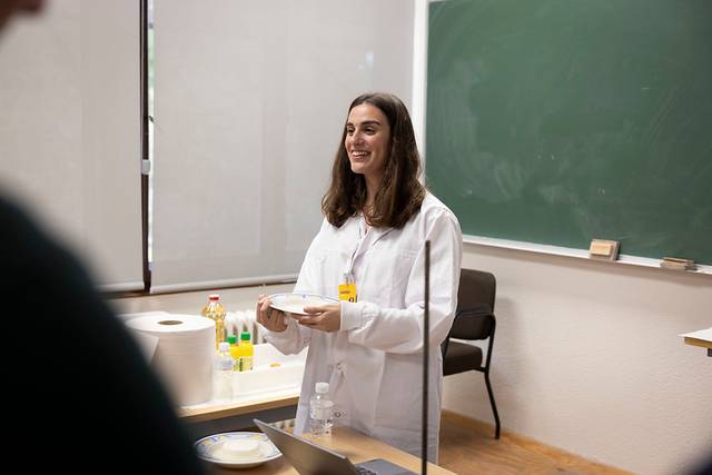 Una mujer sonriente con bata blanca sostiene una placa en un aula.