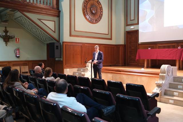 Un hombre habla frente a un grupo de personas en un auditorio con decoración clásica.