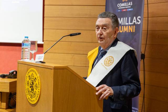 Un hombre habla en un evento académico, vestido con toga y bufanda, frente a un estrado con el logo de la universidad.