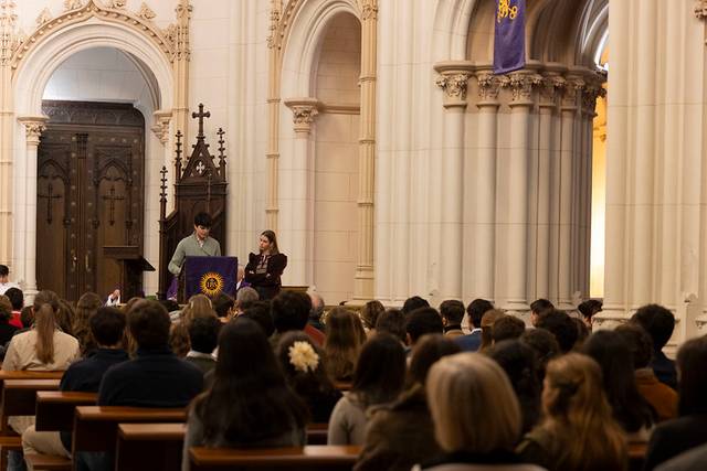 Un grupo de personas asiste a un evento en un entorno religioso con dos presentadores en el escenario.