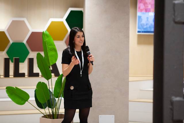 Una mujer presenta en un evento mientras sostiene un micrófono y está rodeada de plantas decorativas.