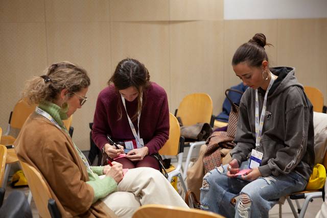 Tres mujeres están sentadas en sillas, concentradas en escribir en cuadernos durante una reunión.