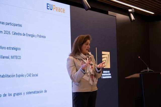 Una mujer está presentando un tema en una conferencia, con una pantalla detrás que muestra información relacionada.