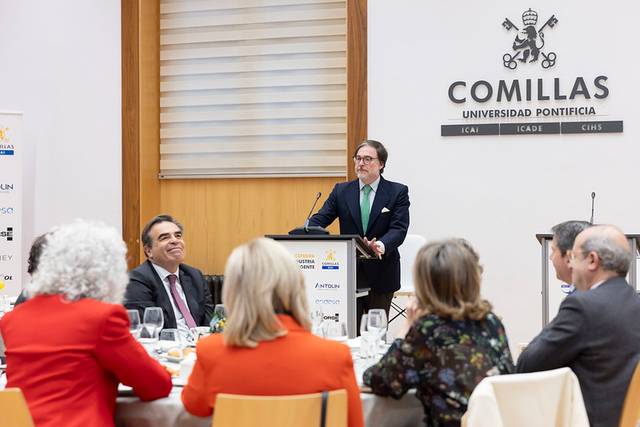Un evento formal en la Universidad Pontificia Comillas donde un orador se dirige a los asistentes en una mesa de banquete.