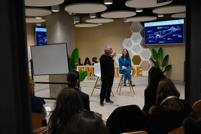Una conferencia en un entorno moderno con un ponente de pie y una mujer sentada, mientras la audiencia observa.