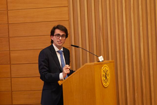 Un joven con gafas habla desde un podio en un entorno formal.