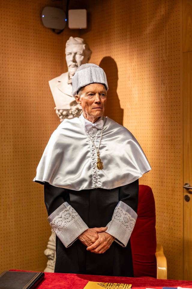 La imagen muestra a un hombre vestido con una toga y birrete, de pie frente a un escritorio en una habitación decorada con una escultura.