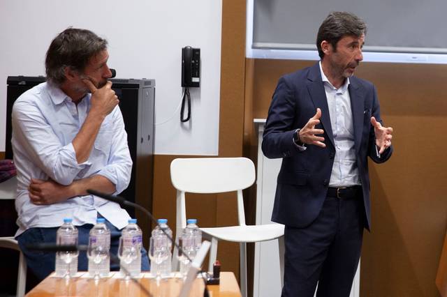 Dos hombres discutiendo en una reunión, uno de ellos gesticulando mientras el otro escucha atentamente.
