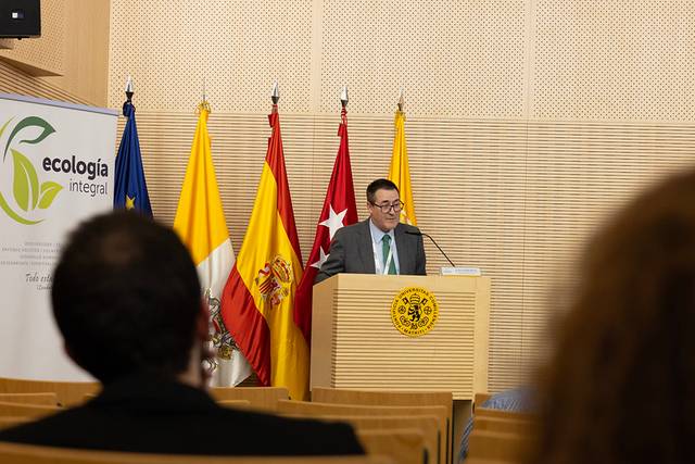 Un presentador está hablando en una conferencia con banderas al fondo.
