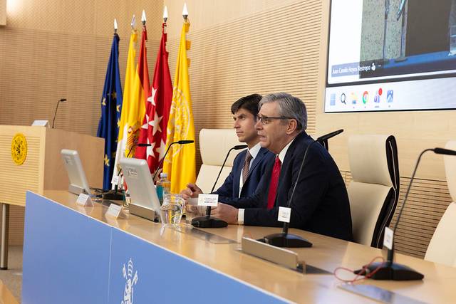 Dos hombres en una conferencia frente a un panel con banderas y una pantalla detrás.