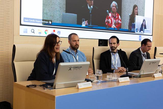 Un panel de discusión con cuatro personas sentadas frente a ordenadores, mientras una conferencia se transmite en la pantalla detrás de ellos.