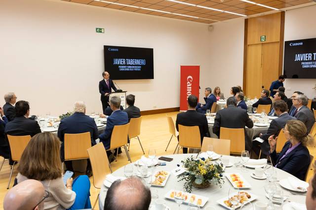 Un hombre está hablando en un evento corporativo con varios asistentes sentados alrededor de una mesa.