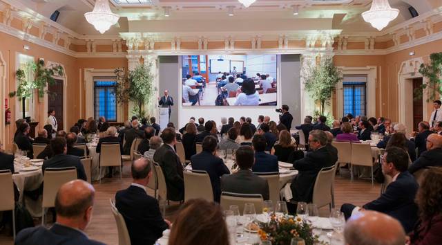 Un evento en un salón elegante con personas sentadas en mesas y una presentación proyectada en una pantalla.
