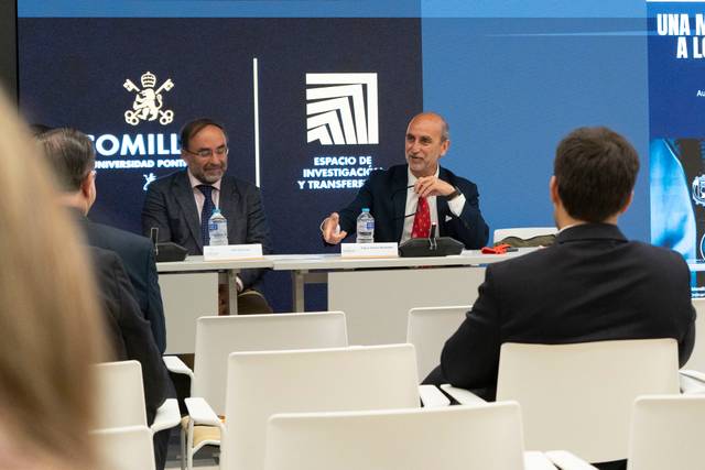Dos hombres están sentados en una mesa durante un evento en un espacio académico.