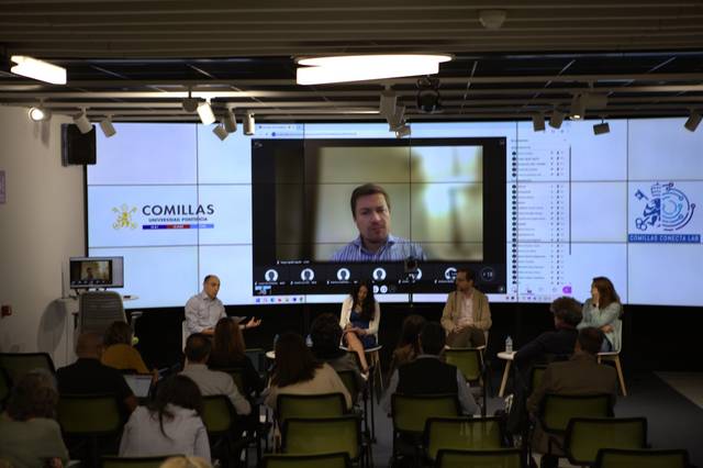 Un panel de discusión en una sala con pantallas y un participante conectado por videoconferencia.