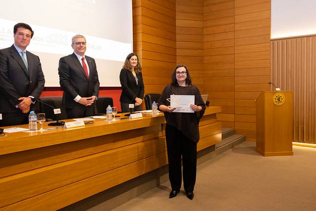 Una mujer posa con un certificado frente a un grupo de personas en un evento formal.