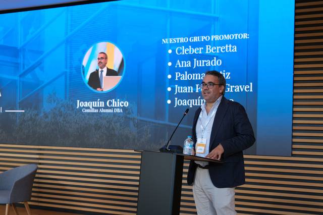 Un presentador habla frente a un proyector en un evento, mostrando un grupo promotor en la pantalla.