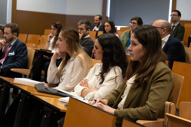 Un grupo de personas asiste a una conferencia en un salón con asientos de madera.