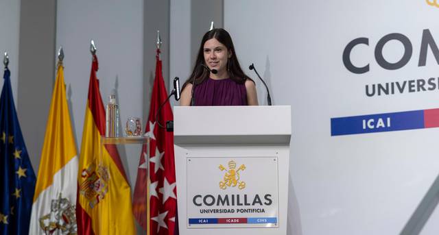 Una mujer de pie en un podio hablando frente a varias banderas y un letrero de la Universidad Pontificia Comillas.