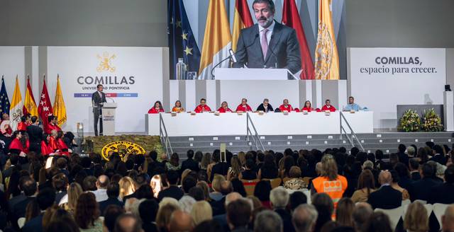 Un evento formal en el que un orador se dirige a una audiencia numerosa en una sala decorada con banderas y un televisor mostrando su imagen.