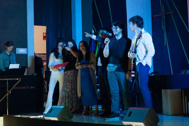 Un grupo de personas en un escenario durante un evento, interactuando y sonriendo mientras uno de ellos sostiene un micrófono.
