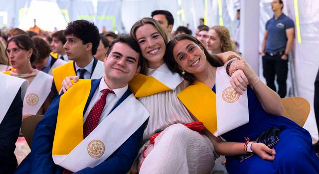 Tres amigos sonríen juntos en un evento de graduación con togas y bandas amarillas.