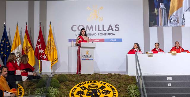 Una mujer habla en un estrado durante un evento en la Universidad Pontificia Comillas, rodeada de banderas y un público atento.