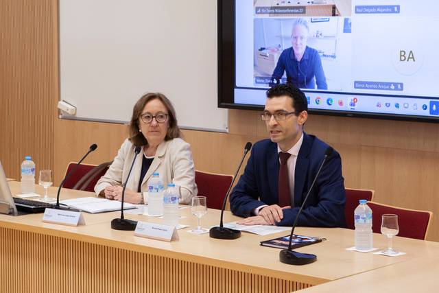 Una presentación formal con dos oradores frente a una audiencia virtual en una sala de conferencias.