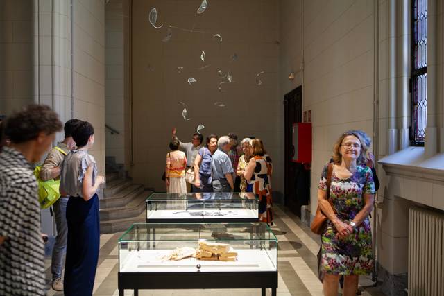 Una exposición en un museo con visitantes observando piezas en vitrinas y instalaciones artísticas en el techo.